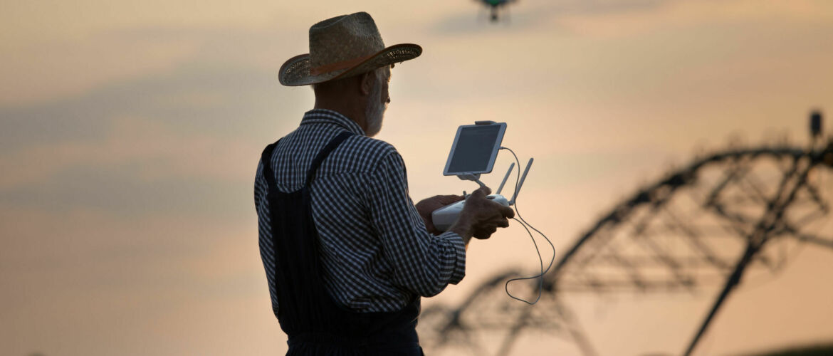 Old farmer with hat holding remote control for drone flying above soybean field with irrigation system in summer